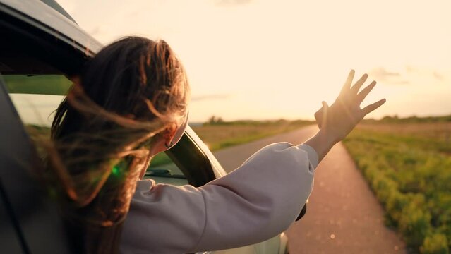 Girl with long hair sits in front seat of car, her hand out window and catching wind, glare of setting sun. Free woman driver rides car catches wind with her hand from car window. Child travels by car