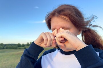 Young woman or teenager scratching itchy and rub eyes with her hands in a park, problems with vision, allergy