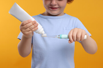 Cute little boy squeezing toothpaste from tube onto electric toothbrush on yellow background, closeup