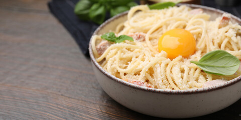 Bowl of tasty pasta Carbonara with basil leaves and egg yolk on wooden table, closeup. Banner design with space for text