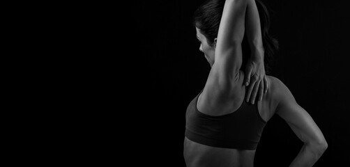 Female sporty muscular woman doing stretching relaxing workout of the shoulders, blades in sport bra, holding the neck the hand on black studio background with empty copy space. Back view. Black