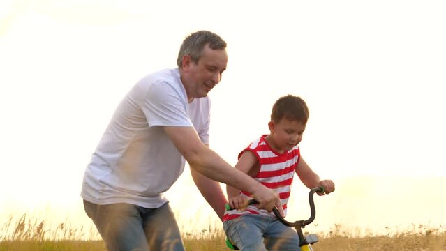 Happy father supports son training to ride bicycle at countryside lit by sunset