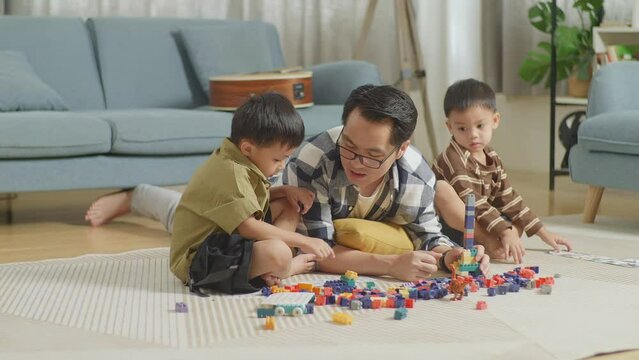 Full Body Of Asian Father And Sons Playing The Construction Set Colorful Plastic Toy Brick At Home. The Kids Assemble Plastic Building Blocks Sitting On A Mat, A Father Lying On The Floor Talking To H