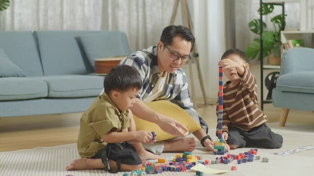 Full Body Of Asian Father And Sons Assemble The Construction Set Colorful Plastic Toy Brick On A Mat At Home
