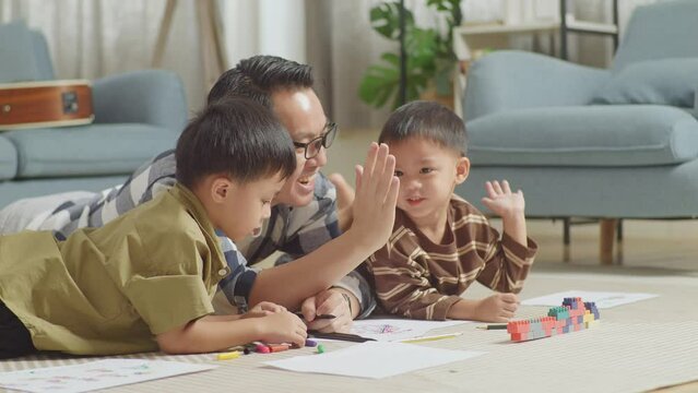Close Up Of Asian Father And Sons On The Floor In The Room With Plastic Toy Brick Giving High Five Celebrating On Success Drawing Together At Home
