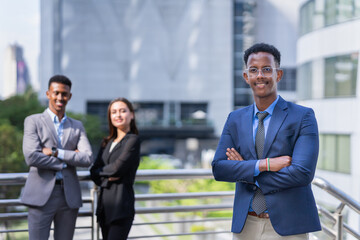 black executive man with glasses in blue suit smilng, standing, cross arm, looking at camera outside building in city. black businessman and caucasian businesswoman standing at background