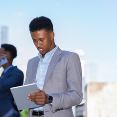 black businessman in grey suit standing and working with tablet outside building in city