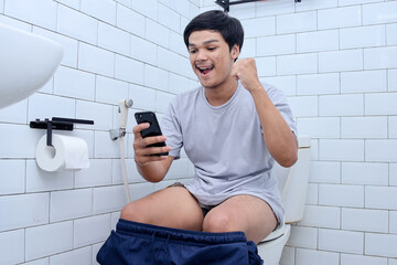 Young man using mobile phone and clenching fist while sitting on toilet bowl in restroom 