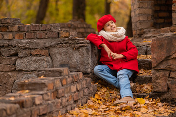 Smiling caucasian girl in a red coat and beret sits on a brick wall on a walk in autumn. 