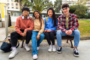 Group of happy and smiling multiracial college student friends looking at camera sitting on park bench. 