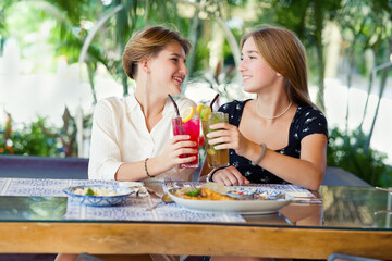 Two happy teenage young girls met each other in the cafe drinking fruit cocktails. Summer warm day on vacations.
