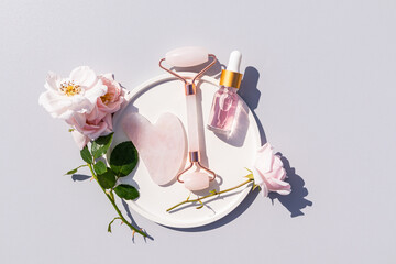 A transparent bottle with a cosmetic product with serum or rose oil lies in a plate with rose petals, a quartz massager, a gua sha scraper. Top view.