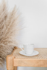 A white coffee cup on a saucer on a rattan bench with pampas gra