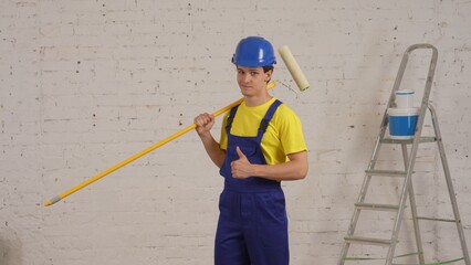 Medium shot of a smiling young worker standing in the room with a roller placed on his shoulder, looking straight at the camera and giving a thumbs up.