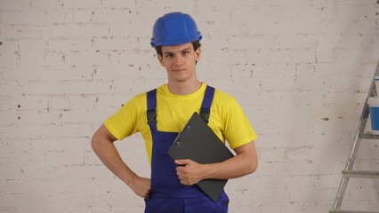 Medium shot of a young construction worker standing in the room, holding a planchette, folder under his arm and looking qt the camera.