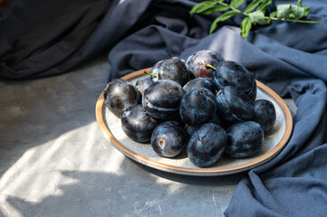 Fresh plums on plate with water drops, dark blue linen tablecloth on gray background with floral natural sunlight shadows. Aesthetic still life