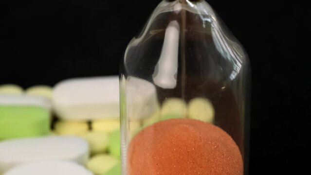 Hourglass and multi-colored pills on black isolated background, drug and tablets. Extending life expectancy through modern medical care.