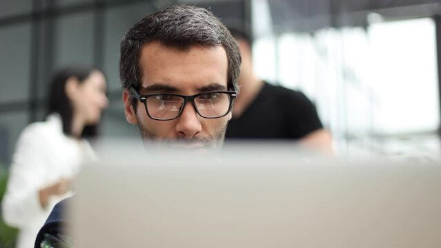 Portrait of handsome bearded man wearing glasses working with laptop in office late, his face lit up by screen
