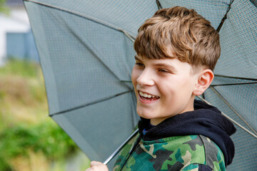 Portrait of a smiling school boy with rainbow umbrella in the park. Kid walking during rain. Happy...