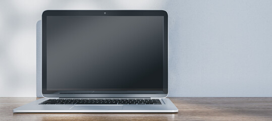 Wide image of laptop with empty mock up screen placed on wooden desk. Concrete wall with shadows in the background. 3D Rendering.