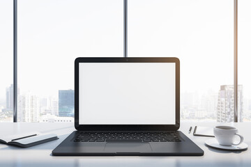 Close up of laptop with empty white mock up monitor, coffee cup and notepad placed on desk. Window with bright city view in the background. 3D Rendering.