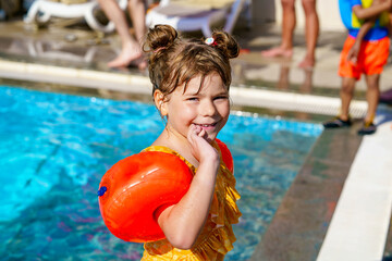 Little preschool girl with protective swimmies playing in outdoor swimming pool by sunset. Child...