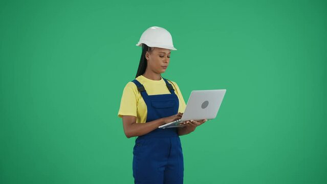 Medium green screen isolated chroma key video of a dark-skinned young female construction worker holding a laptop and typing, inserting data while looking around.