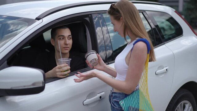 Caucasian Young Man Drinking Juice From Plastic Cup As Woman Entering Offering Glass. Positive Boyfriend Talking With Girlfriend Sitting On Driver's Seat In Car