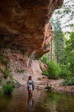 A young woman wades in stream under rock ledge vertical