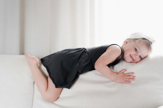 Playful stylish little girl lies astride on white sofa at home