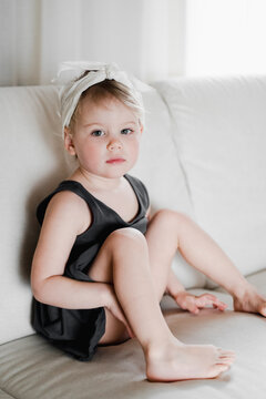 Cute girl 2 years old in a black dress sits on a white sofa