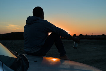 A man sits on the hood of a car in front of the sun at sunset. A man sits conceived with a thermo...