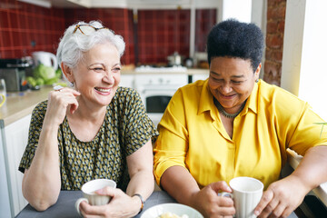 Fototapeta na wymiar Image of two aged female neighbors of caucasian and african ethnicity drinking coffee together in kitchen having fun sharing gossips and rumors, laughing out loud sitting at table