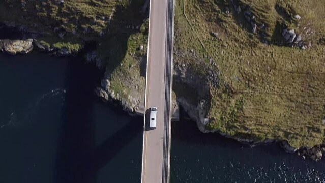 Tracking drone shot of a car crossing the Scalpay island bridge, near the Isle of Harris on the Outer Hebrides of Scotland. The camera tilts to reveal the nearby village, dock and distant mountains.