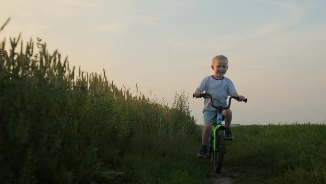 A cheerful, cheerful and cute little boy of 5s old in a striped T-shirt and shorts rides a green-blue bicycle to the camera on a green field during a summer sunset. The boy learns to ride a bike. 4k