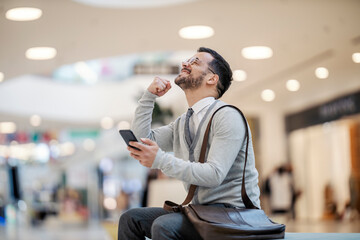 An overjoyed young man is celebrating success while holding his phone and sitting indoors.