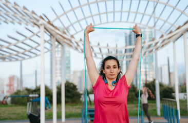 Obraz premium Calm young hispanic woman in sportswear training at sport court using rubber expander looks at camera confidently. Curly caucasian female student at cardio exercise outside. Sport, fitness, health.