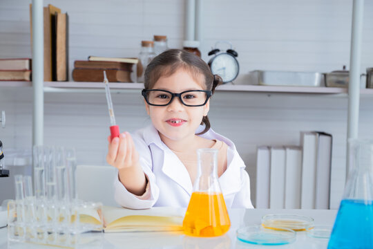 Close-up cute elementary student girl scientist wearing eyeglasses lab coat uniform in lab looking camera hold dropping equipment in laboratory room, lifestyle science kid learning education concept