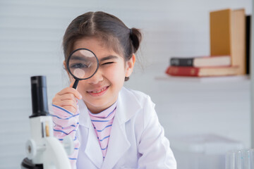 Cute Arabian girl elementary student kid wearing lab coat standing near microscope hold glass...