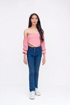 Front view of a slim asian trans woman. Wearing a light pink off-shoulder blouse, jeans and white sneakers. Whole body photo on a white background.