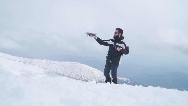Man with beard launches drone from hand while standing in snow high on mountain. Drone taking off for aerial photography or videography flying away.
