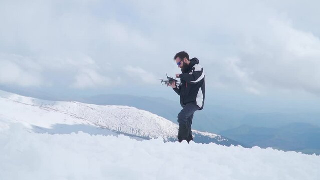 Man standing on snow covered mountaintop holding drone and controller. Aerial photography and videography in extreme climate conditions of winter.