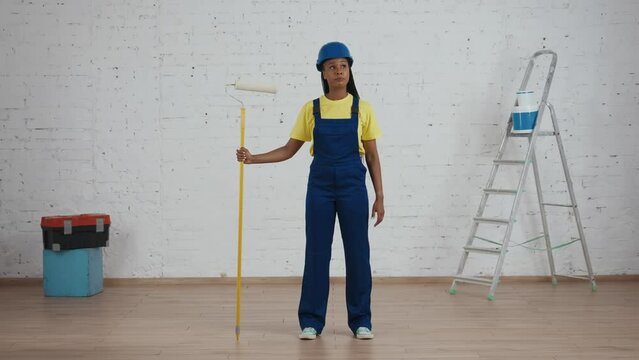 Full length video of a dark-skinned young female construction worker standing in the room with a roller, looking at freshly painted walls and placing her hand on hip.