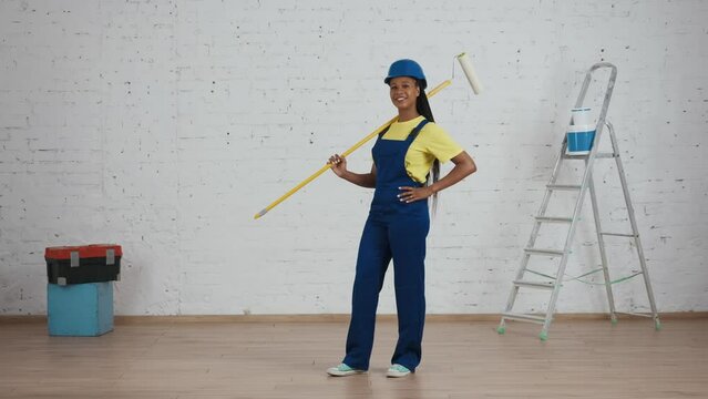 Full length video of a dark-skinned young female construction worker standing in the room with a roller, placing it on her shoulder and looking at freshly painted walls.