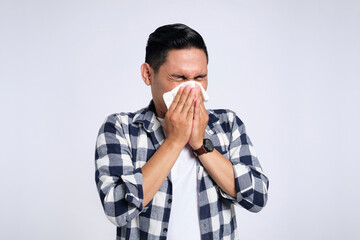 Unhealthy young Asian man in casual shirt blowing nose in paper tissue isolated on white background. Influenza virus concept