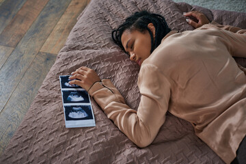 top view of worried african american woman lying on bed near ultrasound, unborn baby, abortion
