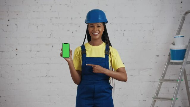 Medium video of a dark-skinned young female construction worker standing in the room under renovation, holding a smartphone with advertising area, workspace mock up and pointing at it.