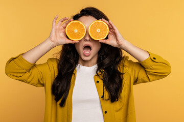 Waist-up portrait of a playful brunette covering the eyes with halves of the citrus fruit on the yellow background. Healthy lifestyle concept