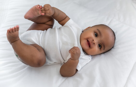 Top view of Happy cute little African newborn baby lying down smiling and looking at camera on white bed in bedroom at home.