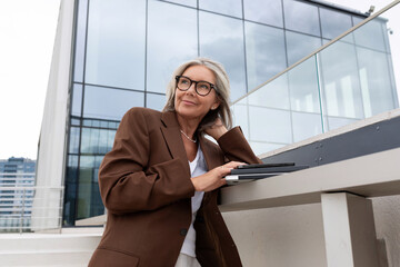 a slender senior business woman with gray hair and glasses dressed in a jacket is waiting for a business meeting standing on the porch